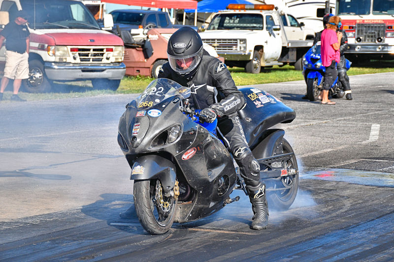 A motorcyclist in a black racing suit and helmet is performing a burnout on a drag racing track, with smoke coming from the rear tire. In the background, several vehicles and spectators are visible.