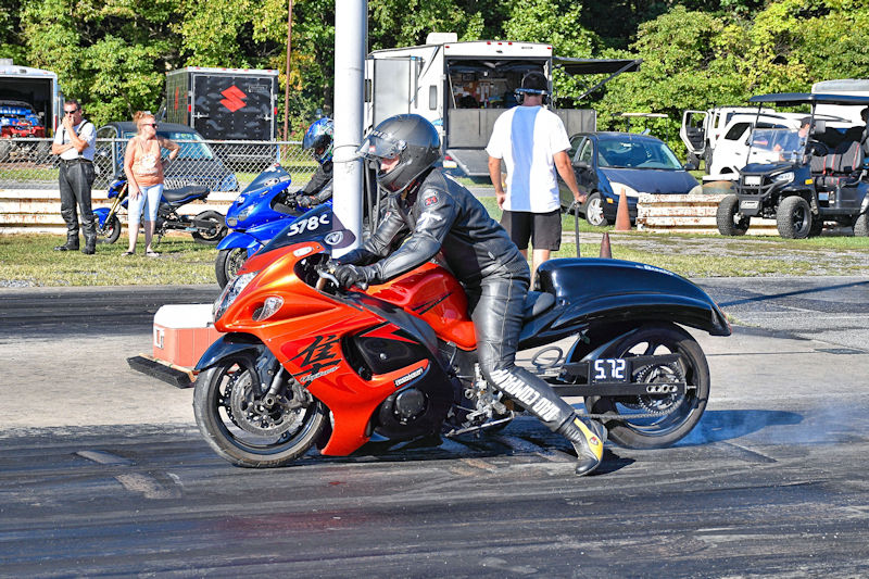 A motorcycle racer in a black and red suit prepares to start a drag race on a track, with smoke coming from the bike's tires. Spectators in the background watch the event.