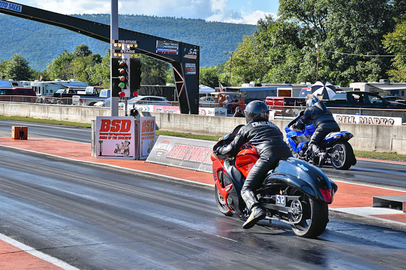 Two motorcycle racers at a drag strip, one in black leather on a red bike and the other in blue attire on a blue bike, preparing to race down the track.