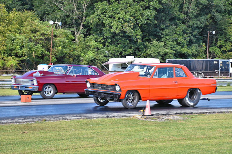 Two classic cars racing on a drag strip, with an orange car lifting its front wheels off the ground and a red car close behind.