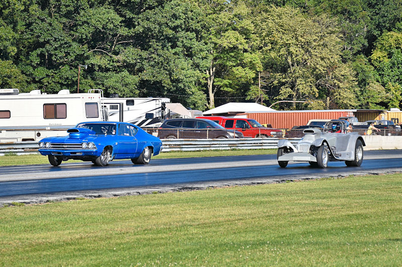 Two racing cars, a blue car and a silver car, speeding down a drag strip with a grassy area and trees in the background.