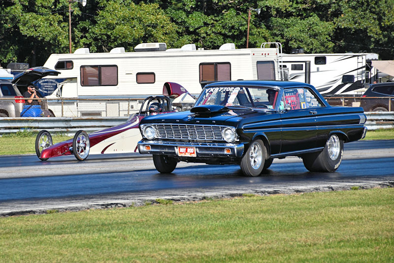 A black classic car and a red dragster racing down a track with spectators and trailers in the background.