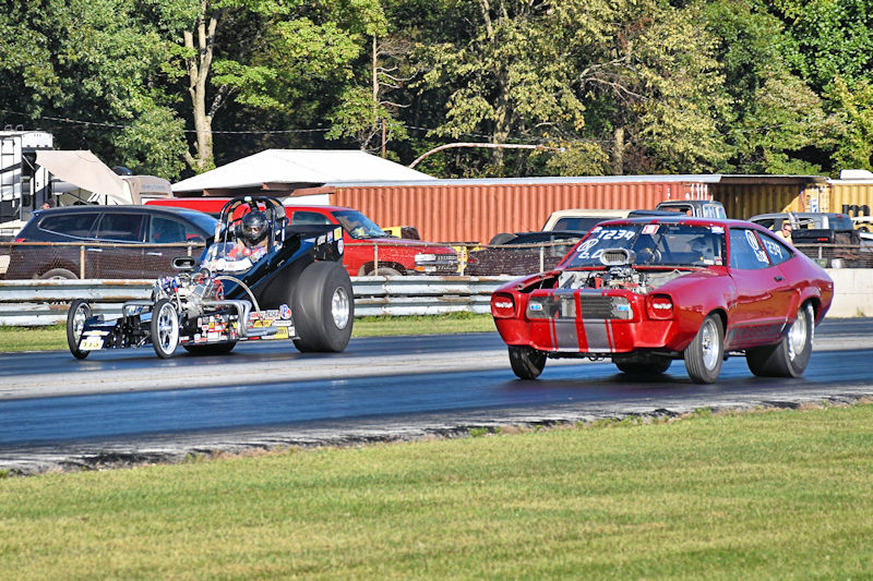 A drag race scene featuring two cars: a black dragster and a red muscle car, both speeding down the track. In the background, there are parked vehicles and trees.
