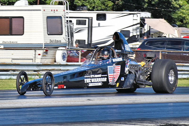 A drag racing car on a track, featuring a driver in a helmet, with a prominent black body and 'It's About The Warrior' slogan on the side.