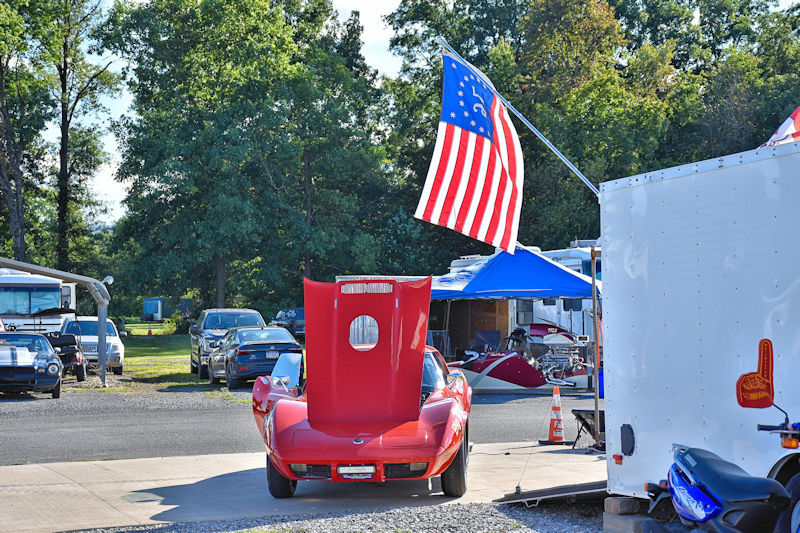 A red sports car with its hood raised is parked in a lot, surrounded by trees and other vehicles. An American flag is flying nearby, with a blue tent set up in the background.