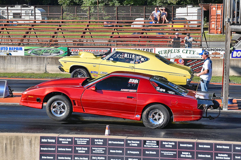 A red drag racing car lifting its front wheels off the ground during a race against a yellow car on a drag strip.