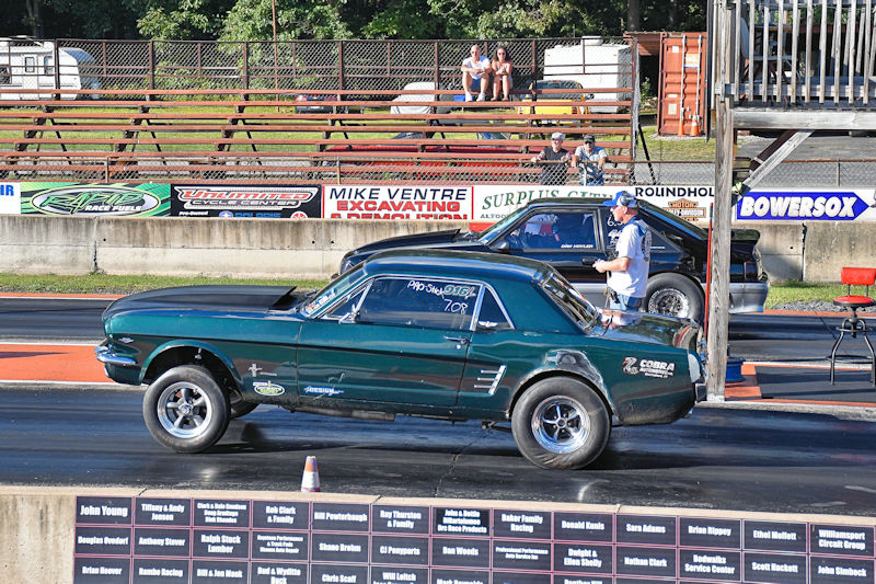 A green classic Mustang car starting a drag race on an asphalt track, with spectators in the background and a black car ready for a race.