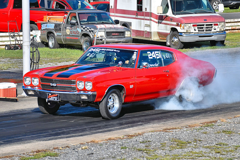 A vibrant red 1970 Chevrolet Chevelle SS is performing a burnout on a drag strip, leaving white smoke from the rear tires. In the background, various vehicles and equipment are visible.