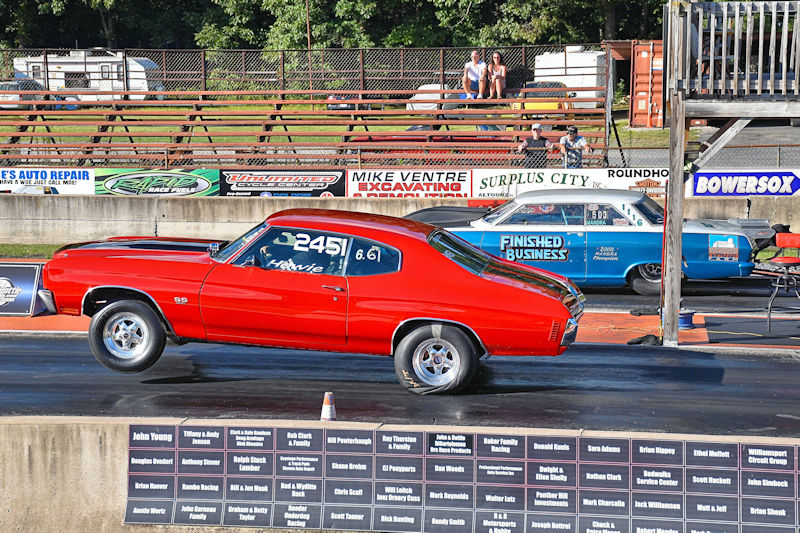 A red muscle car performing a wheelie on a drag racing track, with spectators in the background and a blue car visible in the lane next to it.