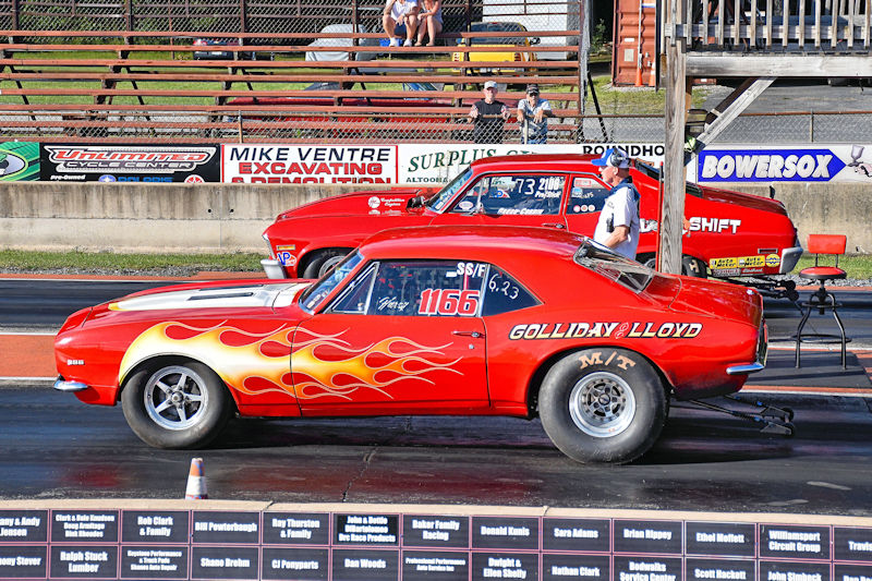A red drag racing car with flame decals is positioned at the starting line next to another car at a race event.