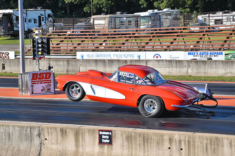 A classic red Corvette race car performing a wheelie on a drag strip during a racing event, with spectators and support vehicles in the background.