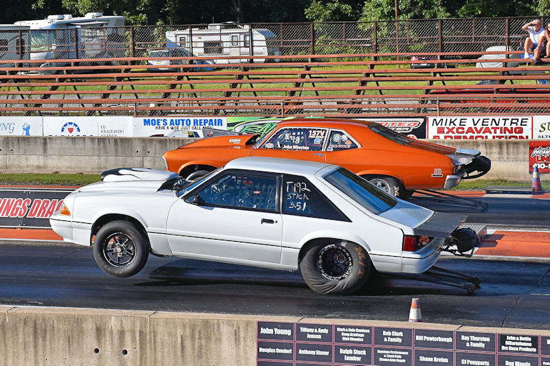 A white race car performing a wheelie on a drag strip, with an orange car visible in the background.