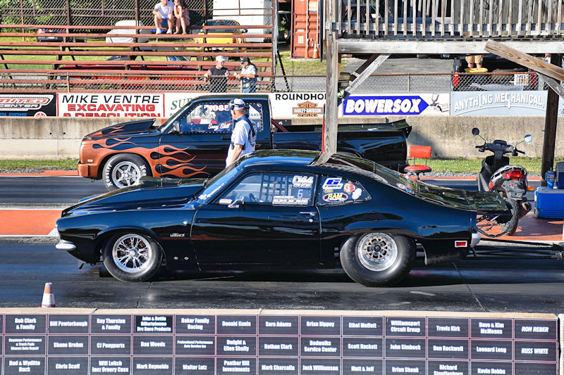 A black race car positioned on a drag strip, with a truck in the background featuring flame decals. A person stands beside the car, preparing for the race.