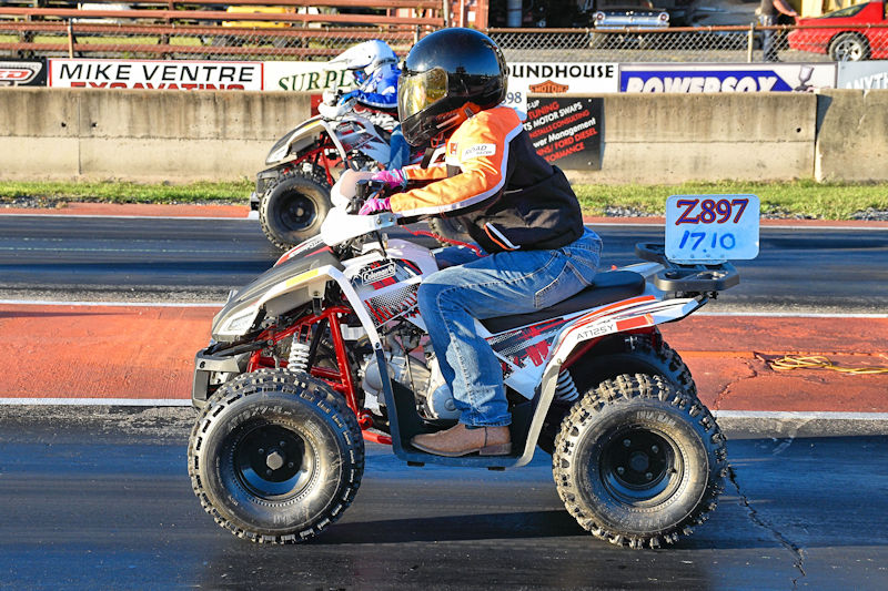 A rider wearing a helmet and protective gear is racing an ATV down a drag strip, with another ATV visible in the background.