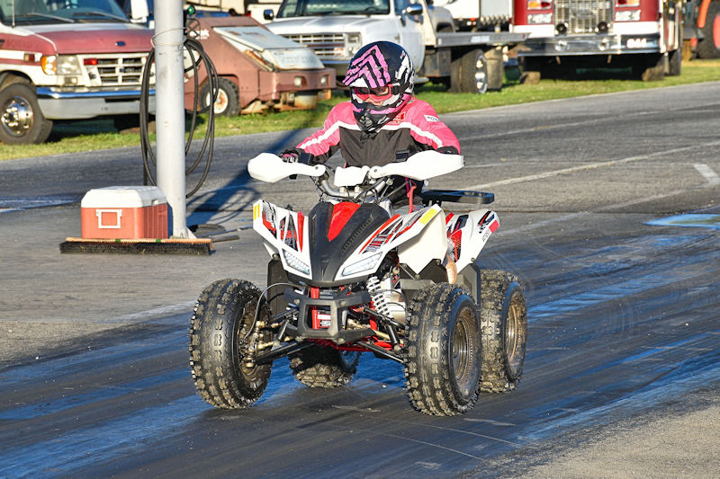 A rider in protective gear riding a four-wheeler ATV on a racing track, with parked trucks in the background.