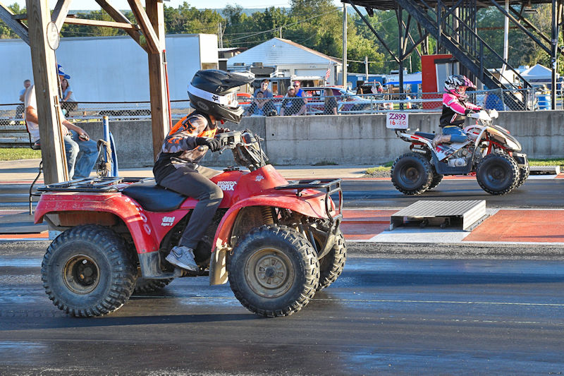 Two young riders racing on ATVs at a drag strip, with a crowd observing in the background.