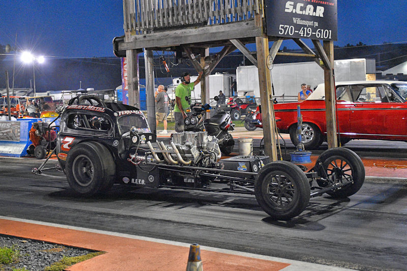 A drag racing car with a tubular chassis and exposed engine is positioned on a racetrack, under a wooden structure with signage. The scene is set at night with several spectators and vehicles in the background.