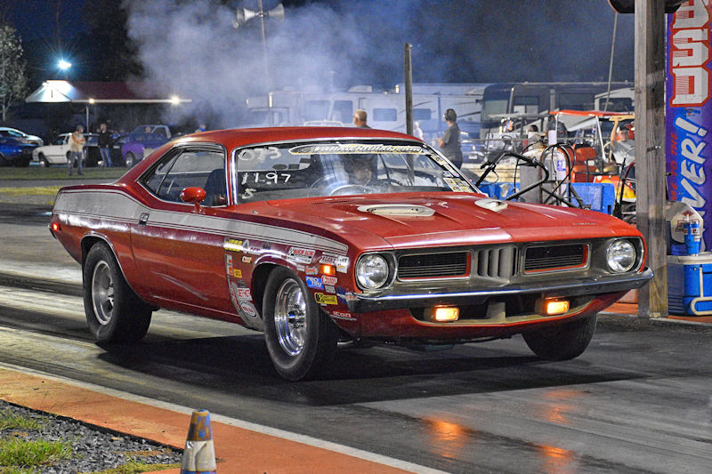 A red classic muscle car, showcasing its racing modifications, is on a drag racing track with smoke rising from the tires, under a night sky with spectators in the background.