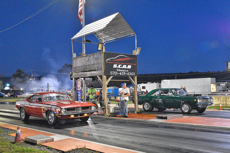 Two cars, one red and one green, lined up at a drag racing track under a night sky, with a timing tower in the background.