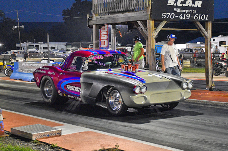 A vintage drag racing car with a colorful paint job, positioned on a racing strip near a grandstand. The car features large rear tires and multiple trophies on the roof, with spectators in the background.