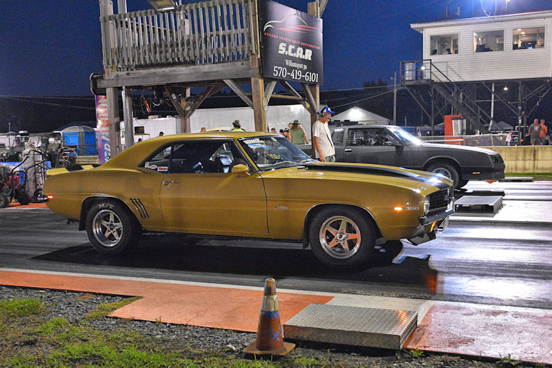 A vintage gold Camaro parked on a drag strip at night, with a timing tower and spectators in the background.
