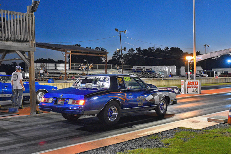 A blue drag racing car prepares to launch down a racetrack at dusk, with spectators in the background and a timing tower visible.