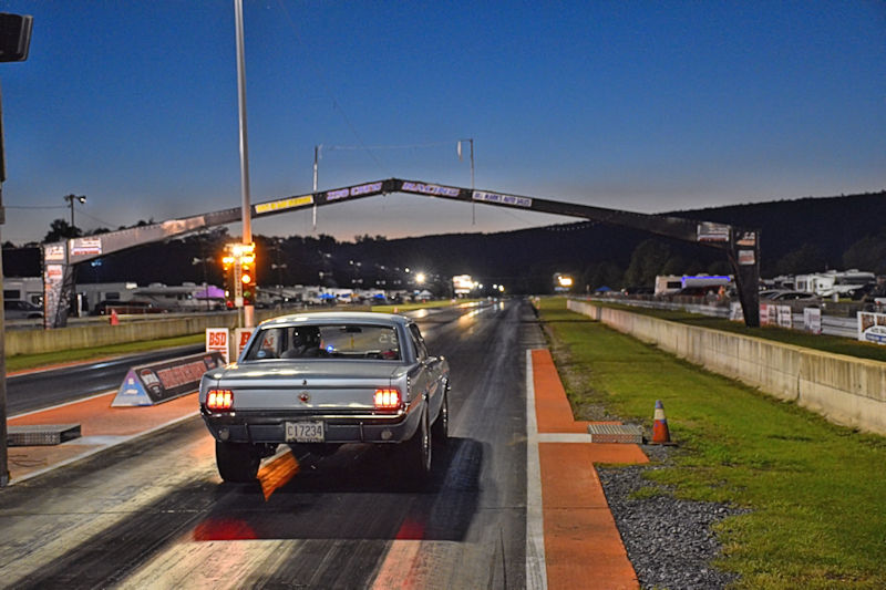 A vintage silver car on a drag racing track at dusk, with a starting line arch and lights illuminated in the background.