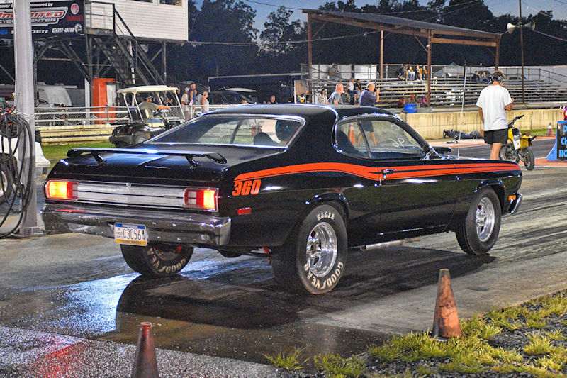 A black muscle car with an orange stripe and '360' emblem, positioned on a drag strip, ready for a race. Spectators and race equipment can be seen in the background under evening lighting.