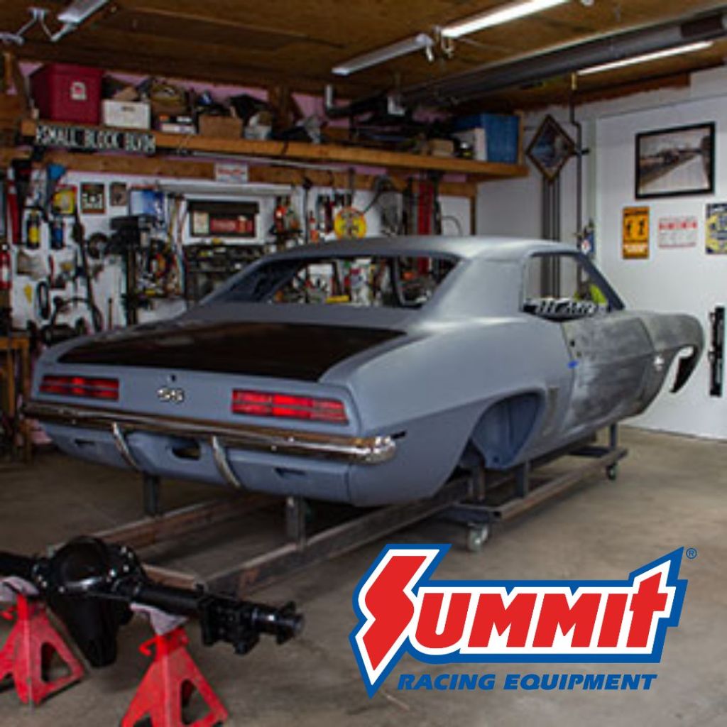 A classic car in a garage, partially restored, with a workbench and tools in the background. The car is mounted on a frame, showcasing its rear with a brown roof and a Summit Racing Equipment logo in the foreground.