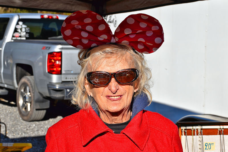 An elderly woman wearing large red and white polka dot Minnie Mouse ears and sunglasses, smiling while standing outdoors in a red coat.