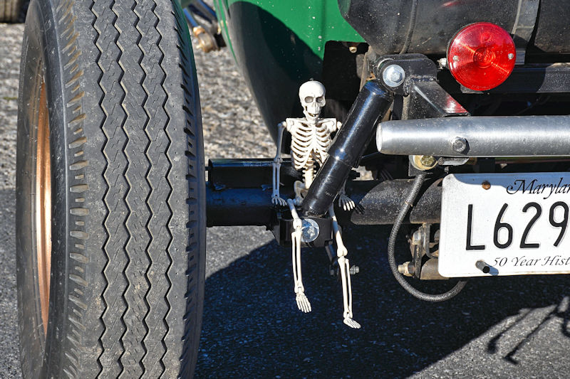 A small skeleton figure sitting on the rear axle of a vintage car, with a close-up view of the tire and license plate.