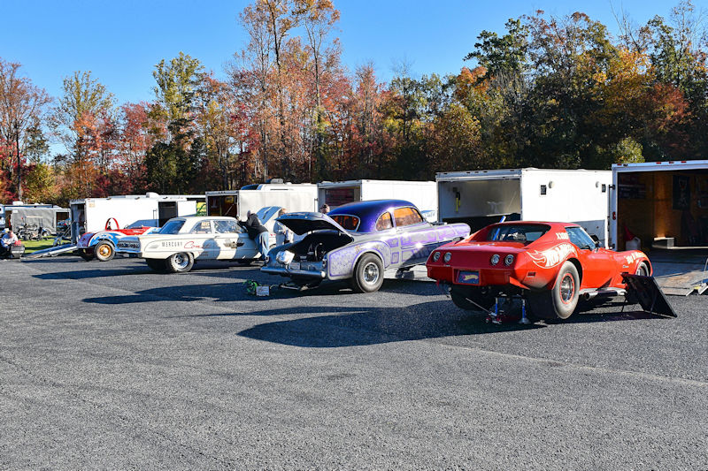 A row of classic cars parked in a gravel lot, with several car trailers in the background. The cars include a purple vintage car with its hood open, a red sports car on jack stands, and other classic vehicles showing mechanical work being done.