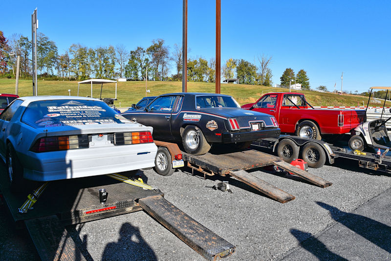 A row of race cars on trailers, including a white car with racing decals, a black car with different sponsors, and a red pickup truck, parked in an outdoor setting with trees in the background.