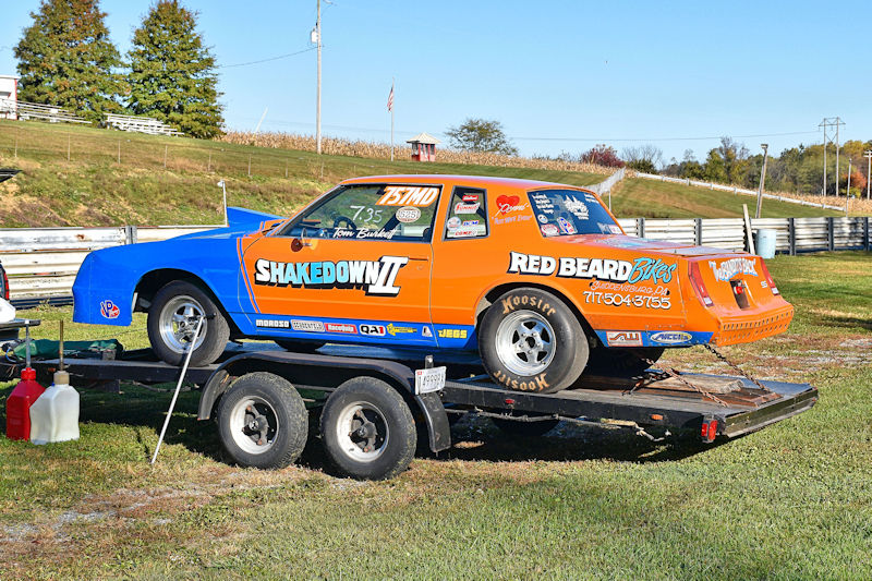 A colorful race car with 'Shakedown II' branding is loaded on a trailer at a racetrack, surrounded by green grass and trees.
