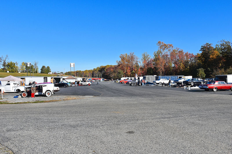A wide view of a parking lot filled with various classic cars and trucks, along with several tents and vendors. Colorful autumn trees are seen in the background under a clear blue sky.