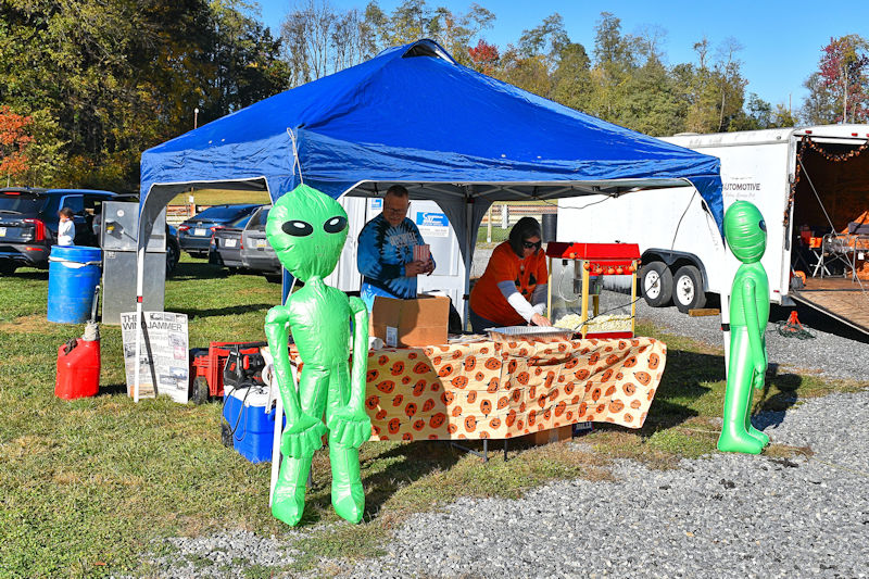 A blue tent at an outdoor event featuring inflatable alien figures, with two people inside preparing food at a table covered with a patterned cloth.