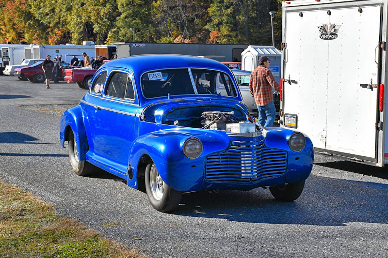 A vintage blue car with an open hood, parked in a racing event area with people and trailers in the background.