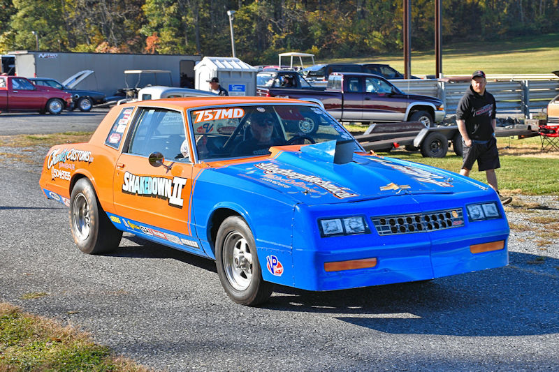 A modified, brightly colored drag racing car with blue and orange paint, featuring the name 'SHAKEDOWN II' on its side, parked on an asphalt track with a man standing nearby.
