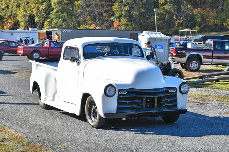 A classic white pickup truck with a simple design is driving in a parking area, surrounded by other vehicles.