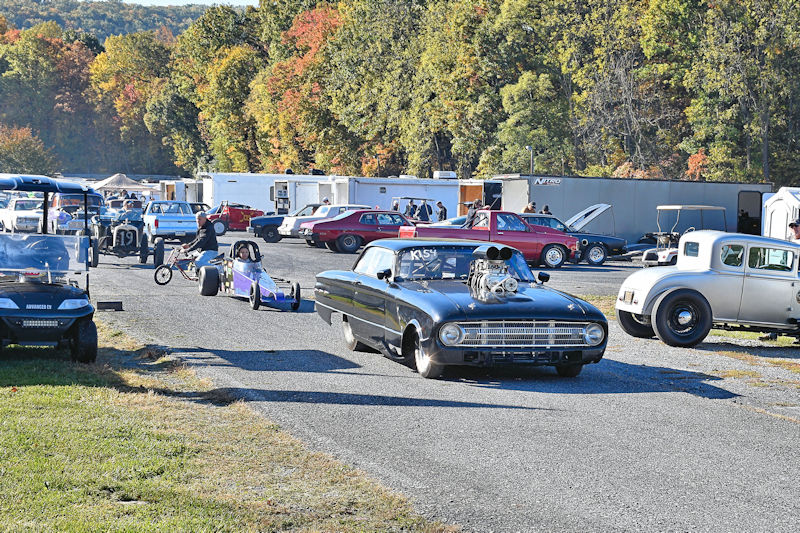 A busy car racing event scene with various vehicles parked and on display, including a black vintage car with a large engine, and a small dragster. There are trailers and other cars in the background, surrounded by autumn trees.