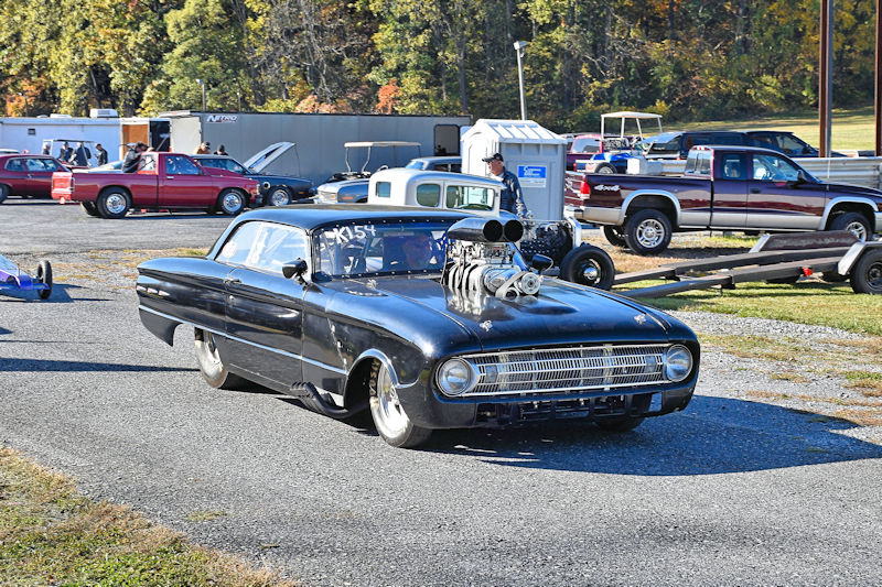 A modified black car with a large supercharger, parked on a racetrack surrounded by various vehicles and trailers.
