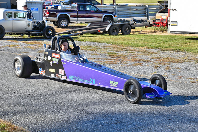 A drag racing car with a purple and black design is driving on a gravel track. The car features various stickers and logos and has a driver seated inside wearing a helmet.