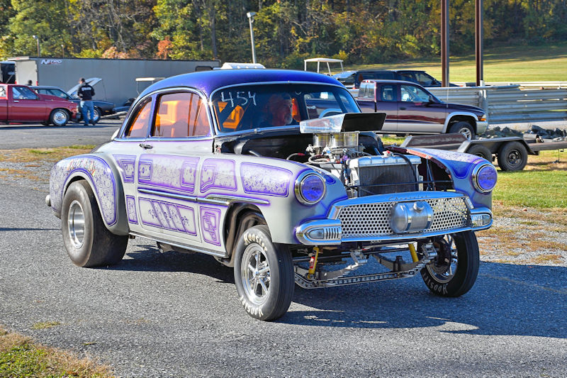 A vintage custom car with a purple and silver paint job and a large engine, parked on a racetrack with other vehicles in the background.