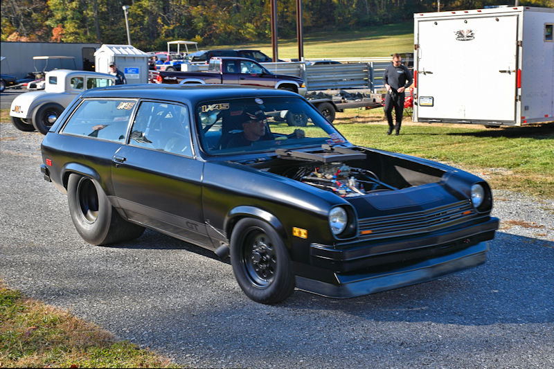 A black modified car with an open hood and racing wheels driving on a gravel road, with a driver wearing a helmet inside. The background features trees and other cars, indicating a racing event.