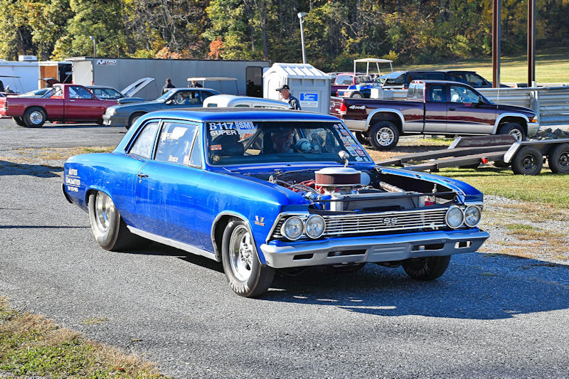 A classic blue 1966 Chevy Chevelle SS with an open hood displaying a large engine, parked at a racetrack with other vehicles in the background.