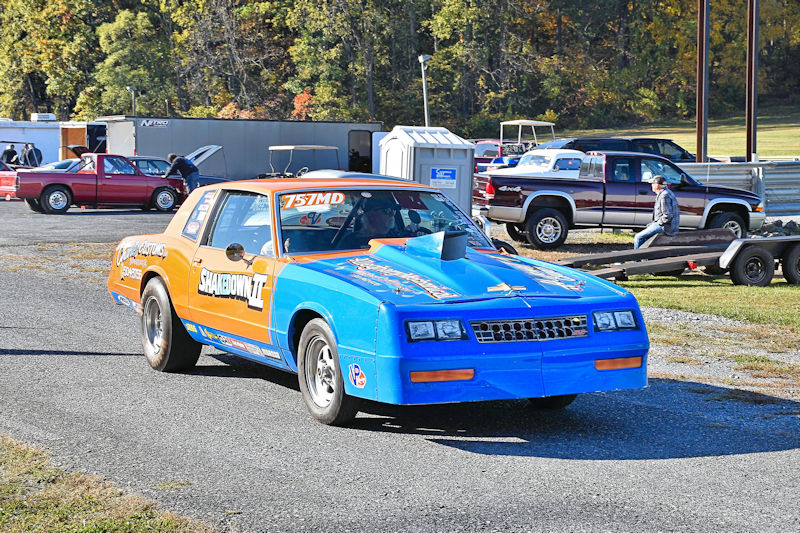 A brightly colored drag racing car with a blue and orange design, parked in a pit area with other vehicles and people in the background.