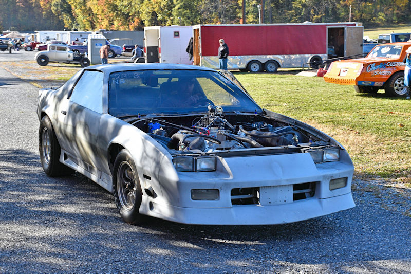 A modified car with an open engine bay, parked on a gravel track surrounded by other vehicles and people in the background.
