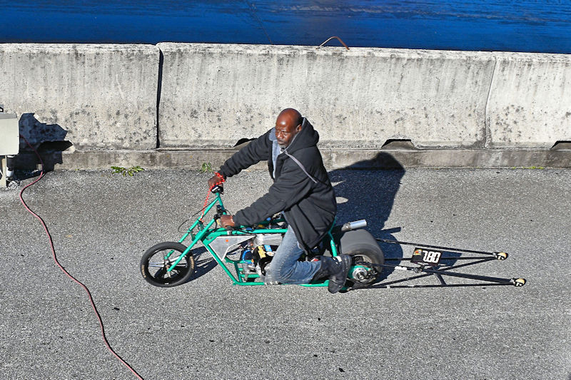 A person riding a small green motorcycle on a concrete surface, with shadows cast by the sun.