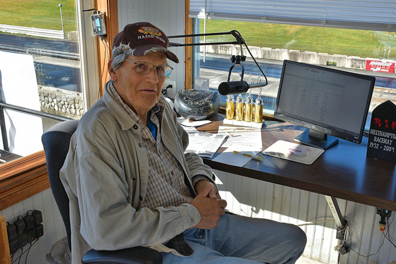 An elderly man sitting at a desk in a raceway control booth, wearing a hat and glasses, with a computer monitor and other equipment on the table.