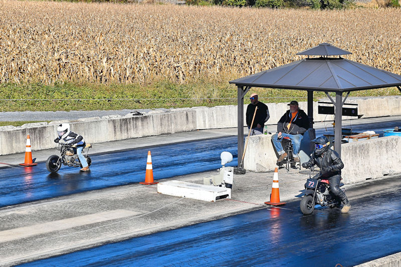 Two riders on small motorcycles racing on a drag strip, with spectators observing from a covered area and cornfields in the background.